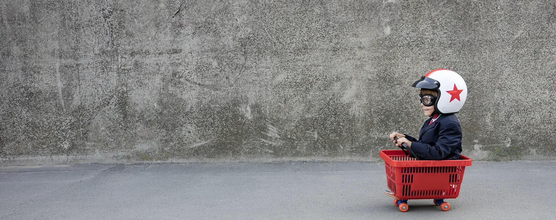 A child in a suit, crash helmet and goggles sitting in a red shopping basket fitted with skateboard wheels, in front of a bare concrete wall.